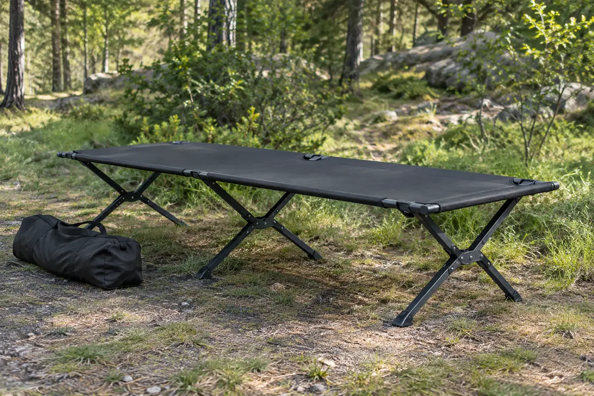 Lit de camp pliable à toile noire et pieds en X installé dans une clairière, avec son sac de transport