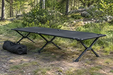Lit de camp pliable à toile noire et pieds en X installé dans une clairière, avec son sac de transport