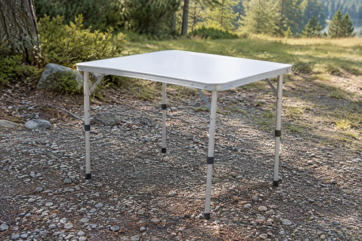 Table de camping pliante blanche avec plateau rectangulaire et pieds réglables en aluminium, installée en extérieur.