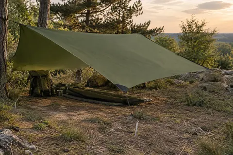 Bâche tarp vert armée tendue dans une clairière, avec cordes et piquets sur un terrain naturel.