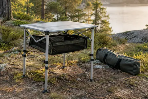 Petite table de camping pliante en aluminium, avec pieds réglables et rangement sous le plateau, installée en clairière.