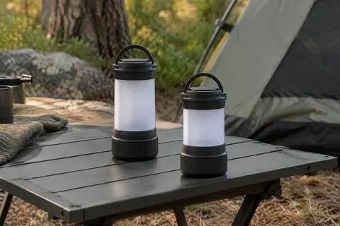 Deux lanternes de camping noires à diffuseur translucide posées sur une table en sous-bois.