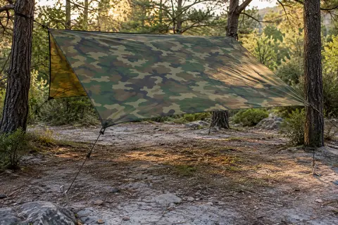 Bâche de camping camouflage tendue dans une clairière, avec ses haubans et ses piquets au sol.