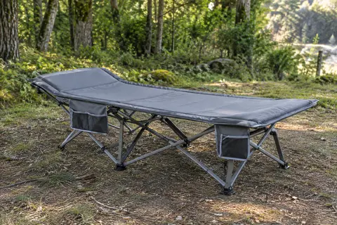 Lit de camp Sportneer gris avec matelas amovible, installé sur un terrain de camping en sous-bois.