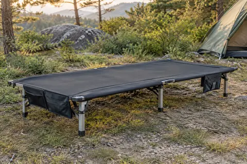 Lit de camp pliant noir avec cadre en aluminium et poches latérales installé dans une clairière.