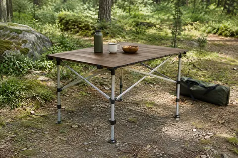 Table de camping en aluminium marron à plateau enroulable, installée dans une clairière avec son sac de transport