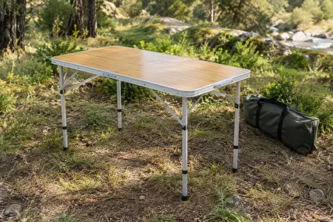 Table pliante de camping en aluminium, finition ton bambou, installée dans une clairière avec son sac