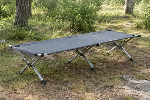 Lit de camp pliant gris à structure métallique installé dans une clairière, avec sa toile tendue.
