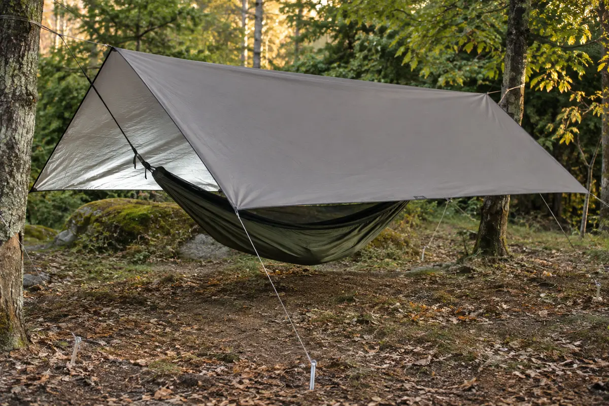 Bâche de camping grise tendue au-dessus d’un hamac dans une clairière, avec cordes et piquets visibles.