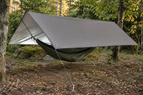 Bâche de camping grise tendue au-dessus d’un hamac dans une clairière, avec cordes et piquets visibles.