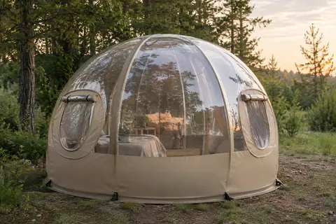 Tente bulle transparente beige installée dans une clairière, avec fenêtres latérales enroulables et vue sur le sous-bois.